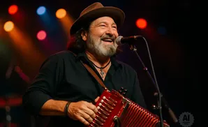 A man with a beard and a hat plays a red accordion on stage with colorful lights.