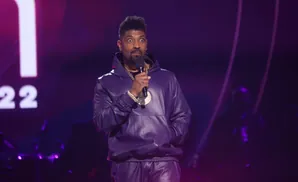 Comedian Deon Cole on stage in a purple leather tracksuit and gold chains.