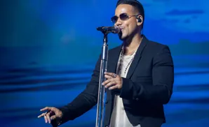 A man in a white shirt and pants sings into a microphone on a dimly lit stage with purple lights.