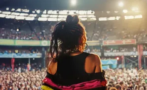 A woman with braided hair and hoop earrings performs on stage in front of a large, cheering crowd in a stadium.