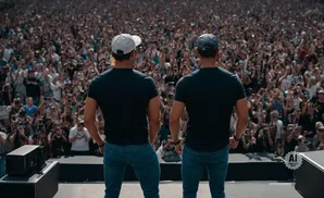 Two men in black t-shirts and jeans stand on a stage facing a large cheering crowd.