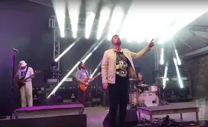 A band performs on stage with bright lights. The lead singer points up.