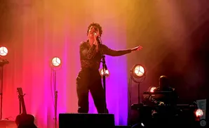 Singer on stage with purple and orange lighting, a guitar and keyboard player in the background.