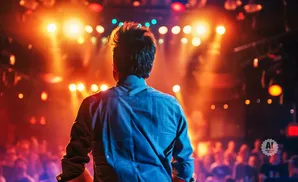 Back of a person in a blue shirt facing a stage with bright orange and yellow lights and a blurred crowd.