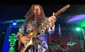 A man with long curly hair plays a guitar on stage, wearing a colorful patterned shirt.