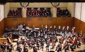 An orchestra performs on stage with a choir in the background, decorated for the holidays.