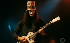 Musician wearing a white mask and a KFC bucket hat plays a white electric guitar on stage.