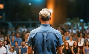 Man in a denim shirt facing away from the camera, addressing a blurry audience.