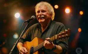 An older man with a mustache smiles as he plays an acoustic guitar and sings into a microphone on stage.