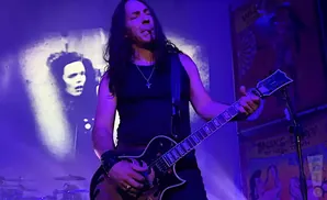 A man with long dark hair plays a black electric guitar, smoking a cigarette, on a dimly lit stage.
