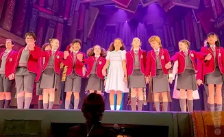 Children in school uniforms on stage, with a bookshelf backdrop.