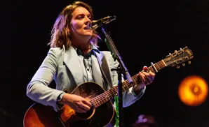 A woman sings and plays an acoustic guitar on stage, lit by stage lights against a dark background.