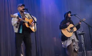 Musicians on stage with blue lighting: a man plays guitar and sings into a microphone, a woman plays guitar.