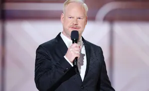 Jim Gaffigan, a comedian, wearing a black suit and white bow tie, holds a microphone and speaks.
