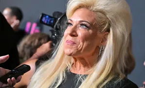 Theresa Caputo with blonde hair, wearing a black dress and sparkling earrings, speaks into a microphone.