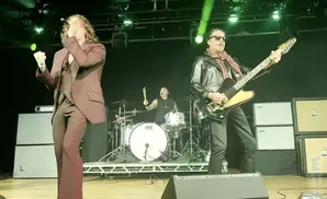 Musicians perform on stage, with a singer in a suit on the left, a guitarist in a leather jacket on the right, and a drummer in the background.