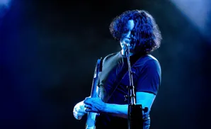 A musician with dark, curly hair plays a guitar under blue stage lights.
