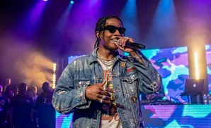 A rapper in a denim jacket and sunglasses performs on stage with purple and yellow lighting.