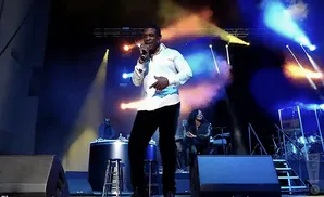 A man in a white shirt and black pants sings into a microphone on a stage with colorful lights.