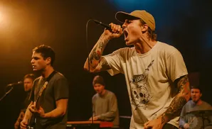 A male singer with a cap and tattoos sings into a microphone on stage, with bandmates behind him.