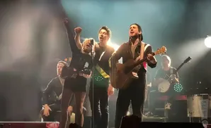 Musicians sing and play instruments on a brightly lit stage, with a female singer in the foreground raising her fist.