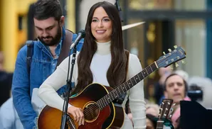 Singer Kacey Musgraves plays an acoustic guitar on stage, wearing a white turtleneck and brown vest.