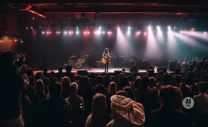 A musician plays guitar on stage at a concert venue, with an audience in the foreground.