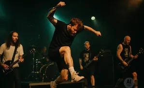Singer leaps mid-song on stage with bandmates playing guitar and drums behind him.