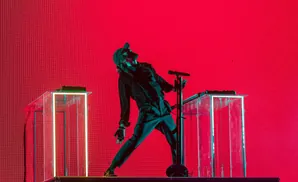 A singer in a black outfit and cap performs on a stage in front of a bright red LED screen.