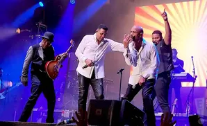 A band performs on stage with bright lights and a large screen behind them.