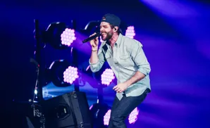 Man in a baseball cap and denim shirt sings into a microphone on stage with purple lights.