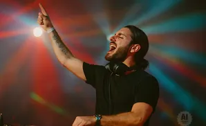 DJ with headphones on and arm raised, silhouetted against colorful stage lights.