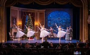 Ballerinas in white tutus dance on a stage with a Christmas tree and a snowy window backdrop, while an orchestra plays in the foreground.
