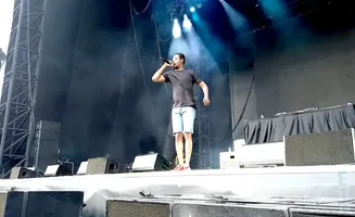 A man in a black t-shirt and denim shorts performs on stage with a microphone.