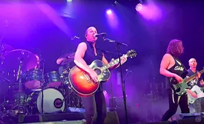 A bald man with a sleeveless shirt plays an acoustic guitar and sings into a microphone on a stage with purple lighting.