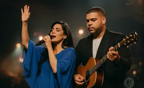 A woman sings into a microphone with her hand raised, a man plays an acoustic guitar beside her.