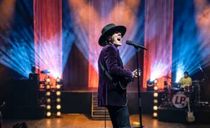 Singer LP performs on stage wearing a black hat and purple velvet jacket, with a microphone and guitar.