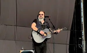 A man with a beard and sunglasses plays an acoustic guitar on stage.