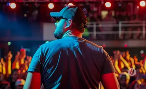 Man in a baseball cap and sunglasses on stage, facing away from the camera, with a cheering crowd in the background.