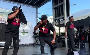 Three men in black hoodies and pants perform on a stage.