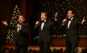 Three men in tuxedos sing into microphones on stage, with a Christmas tree and sparkling lights behind them.