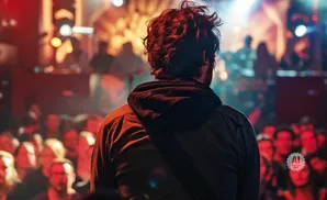 Man with messy red hair from behind watches a band perform on a stage with a blurred audience.