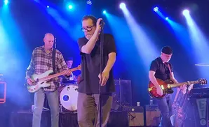 A band performs on stage with blue lights, featuring a singer, bassist, and guitarist.