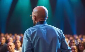 Bald man in a blue shirt giving a speech to a blurred audience under stage lights.