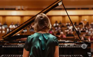 A pianist in a green dress faces a grand piano, with an audience in the blurred background.