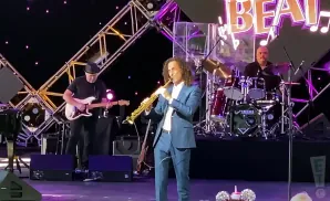 Kenny G plays saxophone center stage with a guitarist and drummer behind him at a concert.