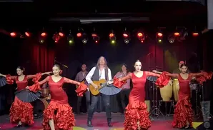 Flamenco dancers in red dresses perform with a guitarist and percussionists on a dimly lit stage.