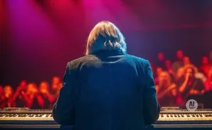 A keyboardist plays on stage with a blurred audience and vibrant pink lighting.