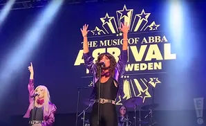 Two female singers perform on stage with a banner reading 