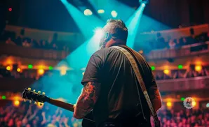 Guitarist with tattoos plays on a brightly lit stage before a cheering audience.